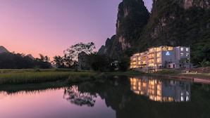Un hotel moderno con varios pisos, ubicado junto a una masa de agua tranquila, rodeado de vegetación exuberante y montañas.