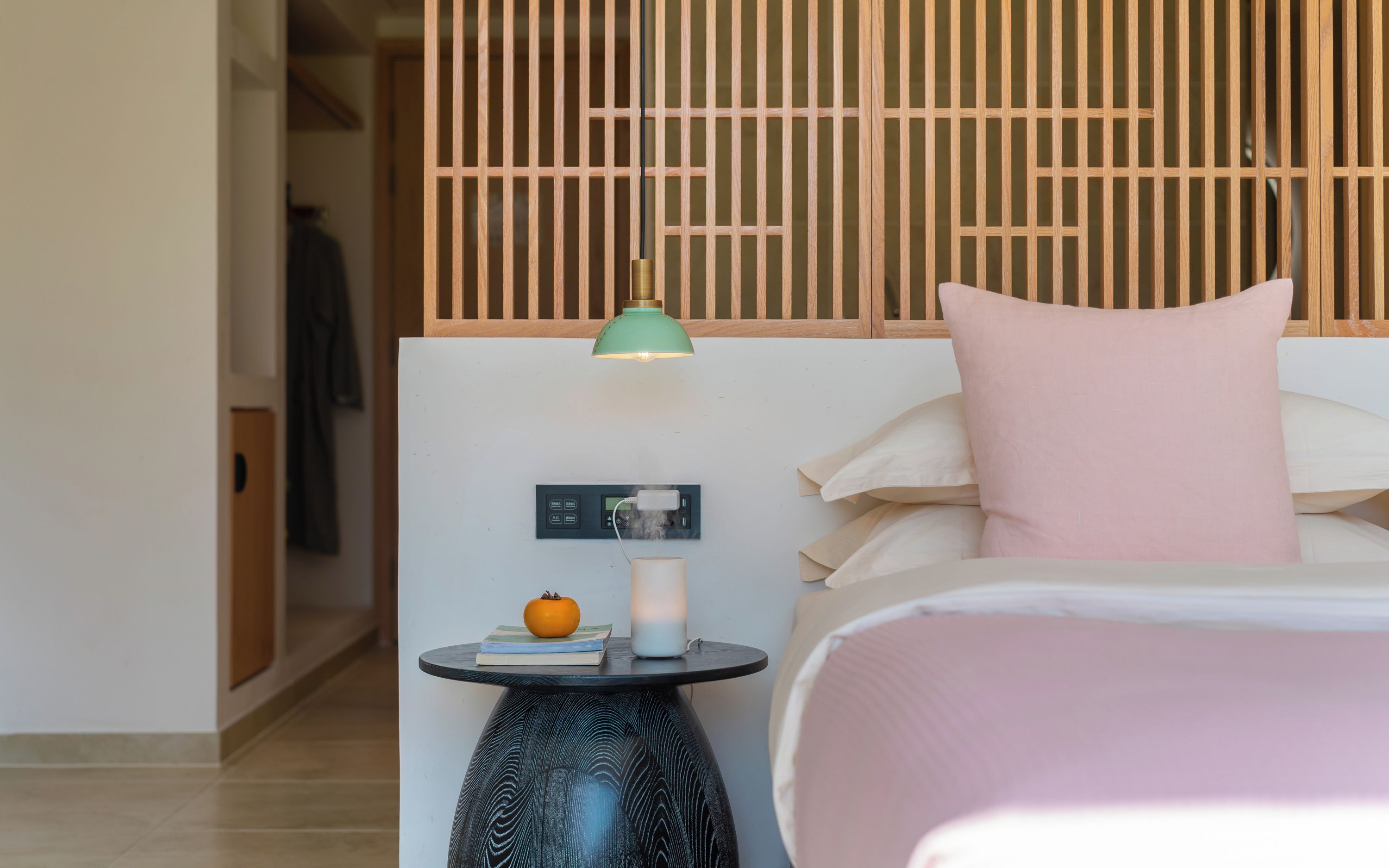Una habitación de hotel con una cama, una mesita de noche con una lámpara, una pequeña mesa con una vela y una manzana, y un panel de control en la pared.