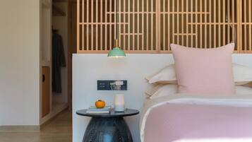 Una habitación de hotel con una cama, una mesita de noche con una lámpara, una pequeña mesa con una vela y una manzana, y un panel de control en la pared.