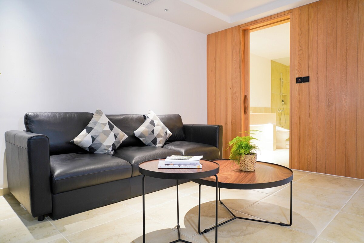 A modern living room with a leather sofa, two patterned throw pillows, a small round coffee table with books, and a potted plant.
