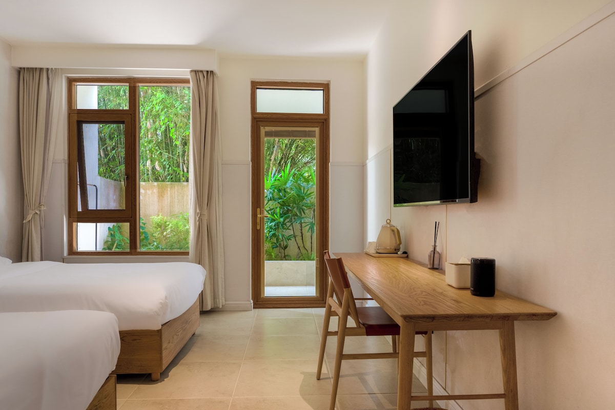 A hotel room with two beds, a desk, a TV, and a view of greenery outside.