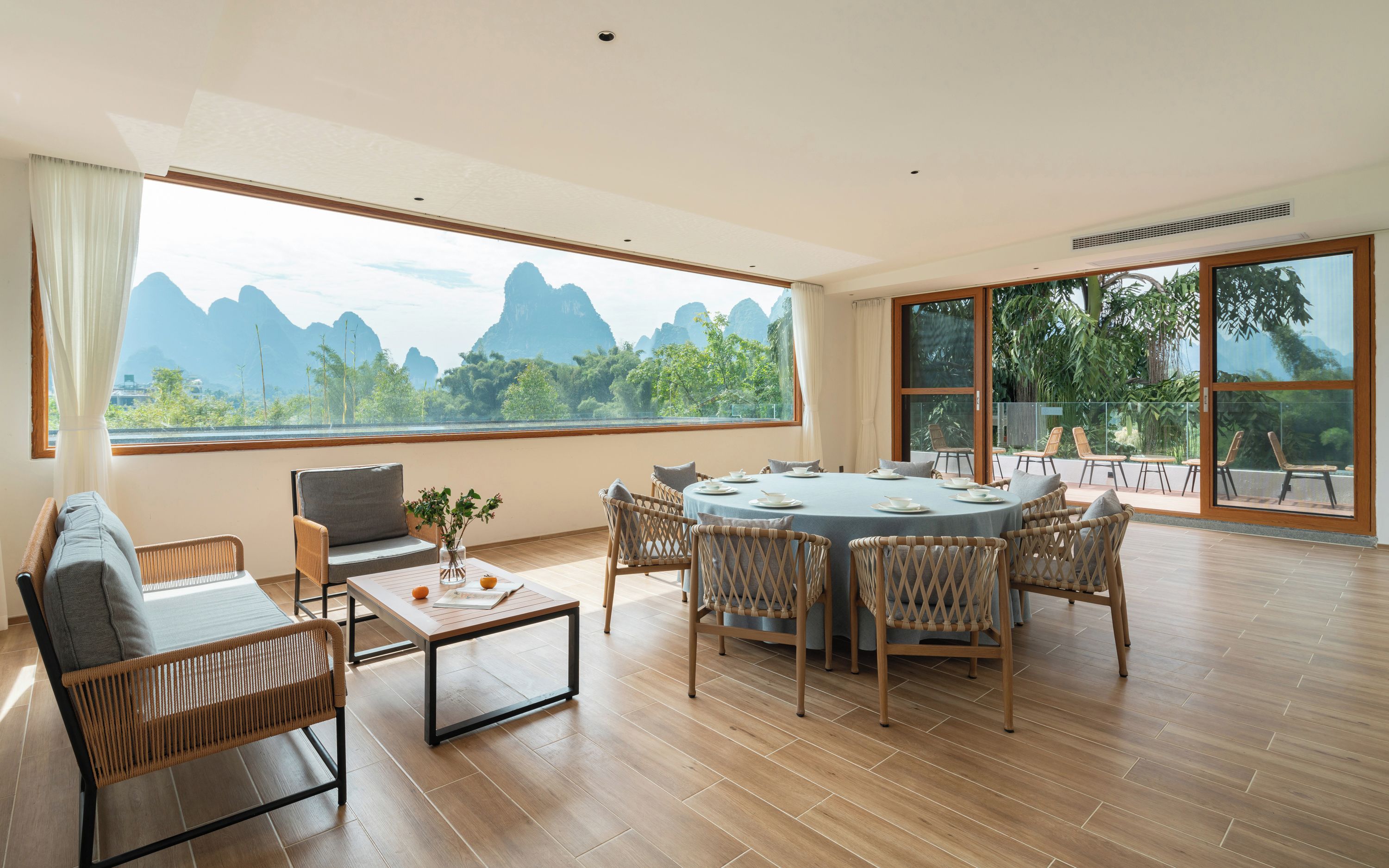 Una habitación amplia con una gran mesa de comedor rodeada de sillas, un sofá, y un amplio ventanal con vistas a montañas y vegetación.
