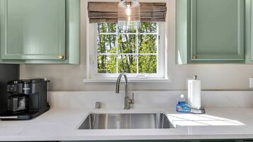 A kitchen with a stainless steel sink, a coffee maker, a window with a view of trees, and a roll of paper towels.
