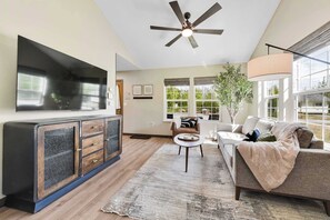 A modern living room with a flat-screen TV, a ceiling fan, a sofa, a coffee table, and a large window with a view of greenery.