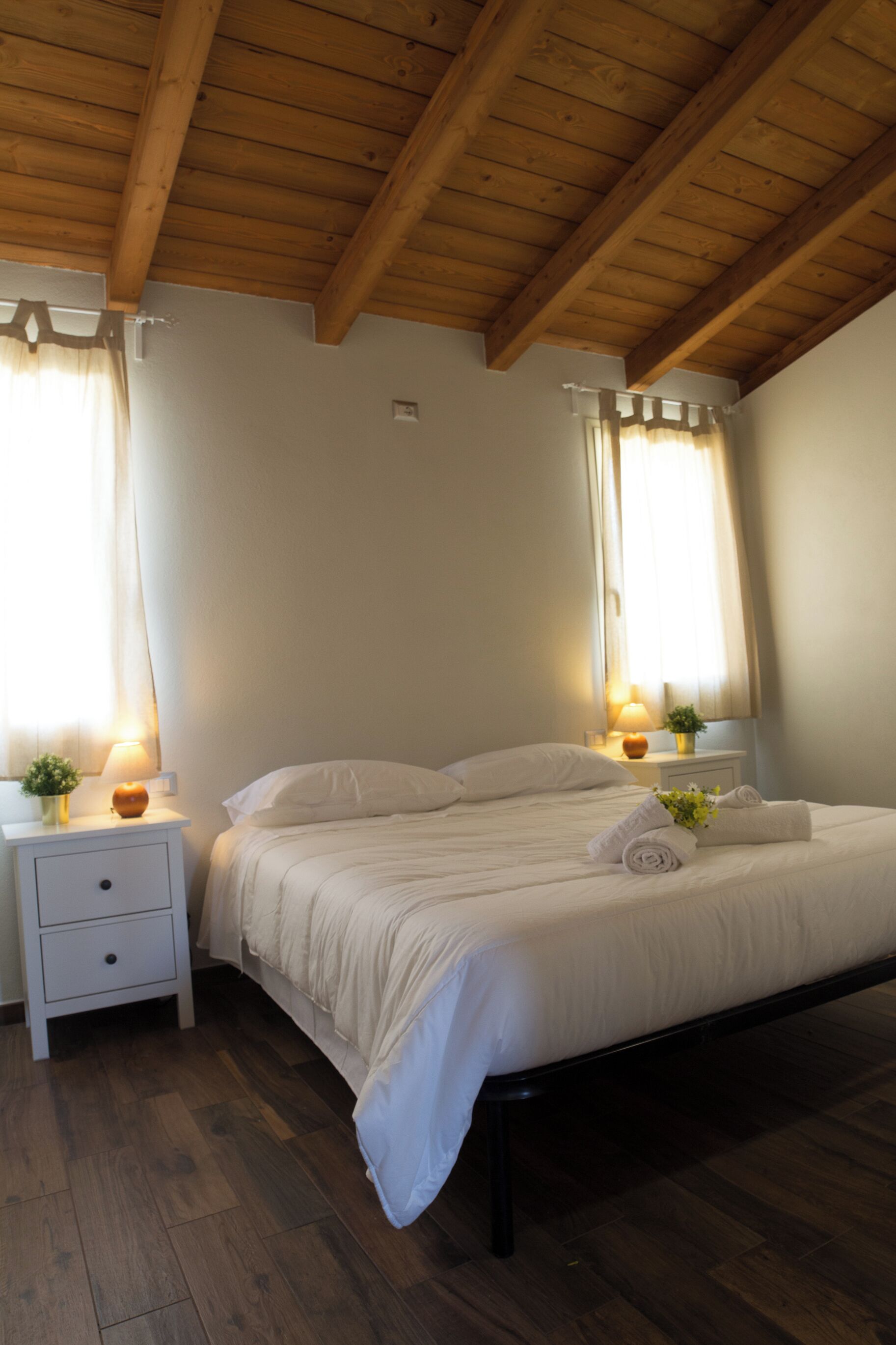 Un dormitorio con techo de madera, una cama con ropa blanca, dos mesitas de noche con lámparas y una planta pequeña en cada una.