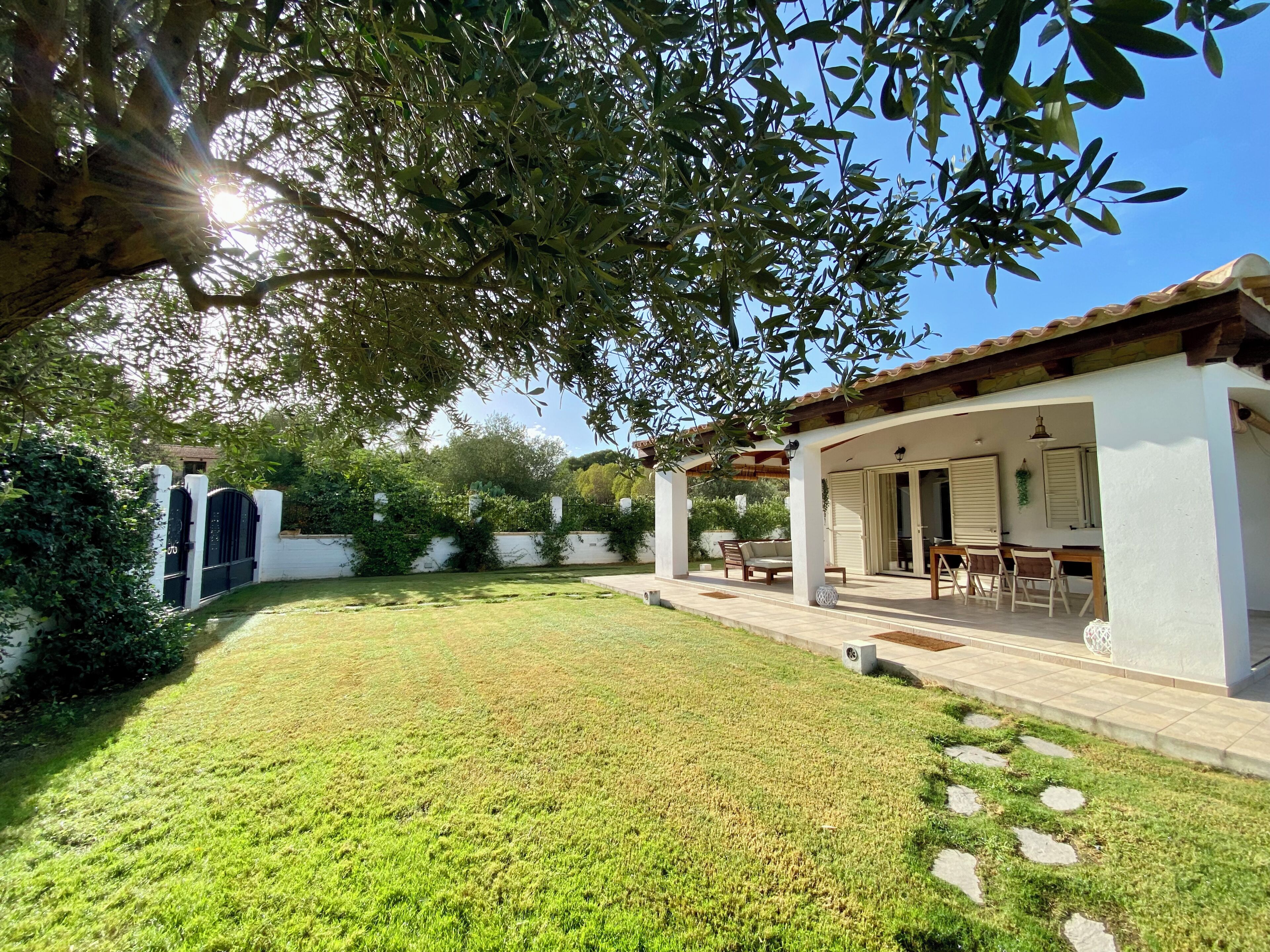 Un'area salotto esterna coperta con tavolo e sedie da patio, una parete bianca con una finestra e un giardino con prato e sentiero acciottolato.