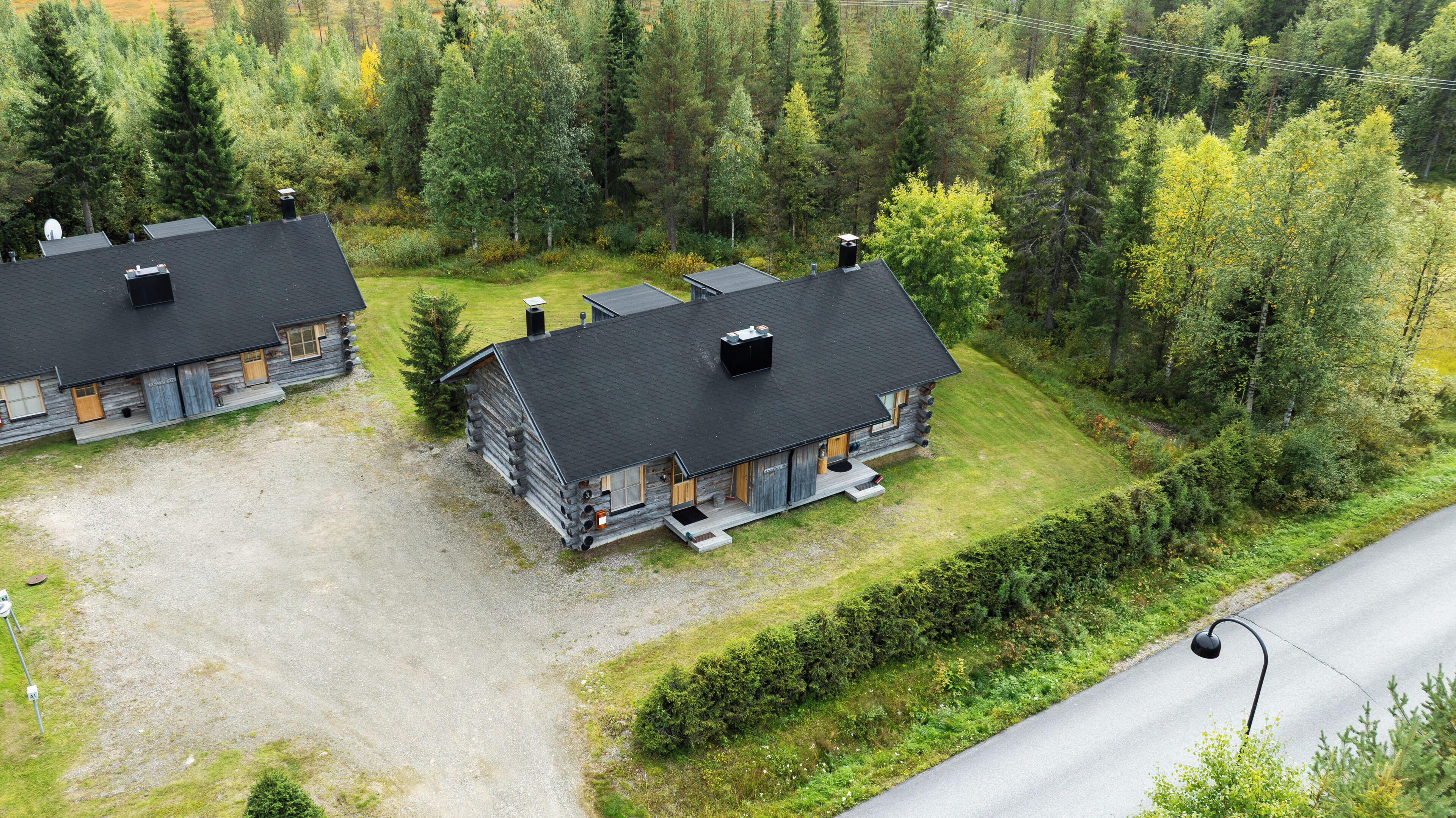 A lodge with two cabins, each featuring a black roof and log construction, surrounded by dense forest.