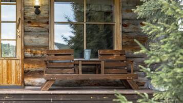 A wooden cabin with a bench and a table, a window with a view of trees, and a light fixture on the wall.