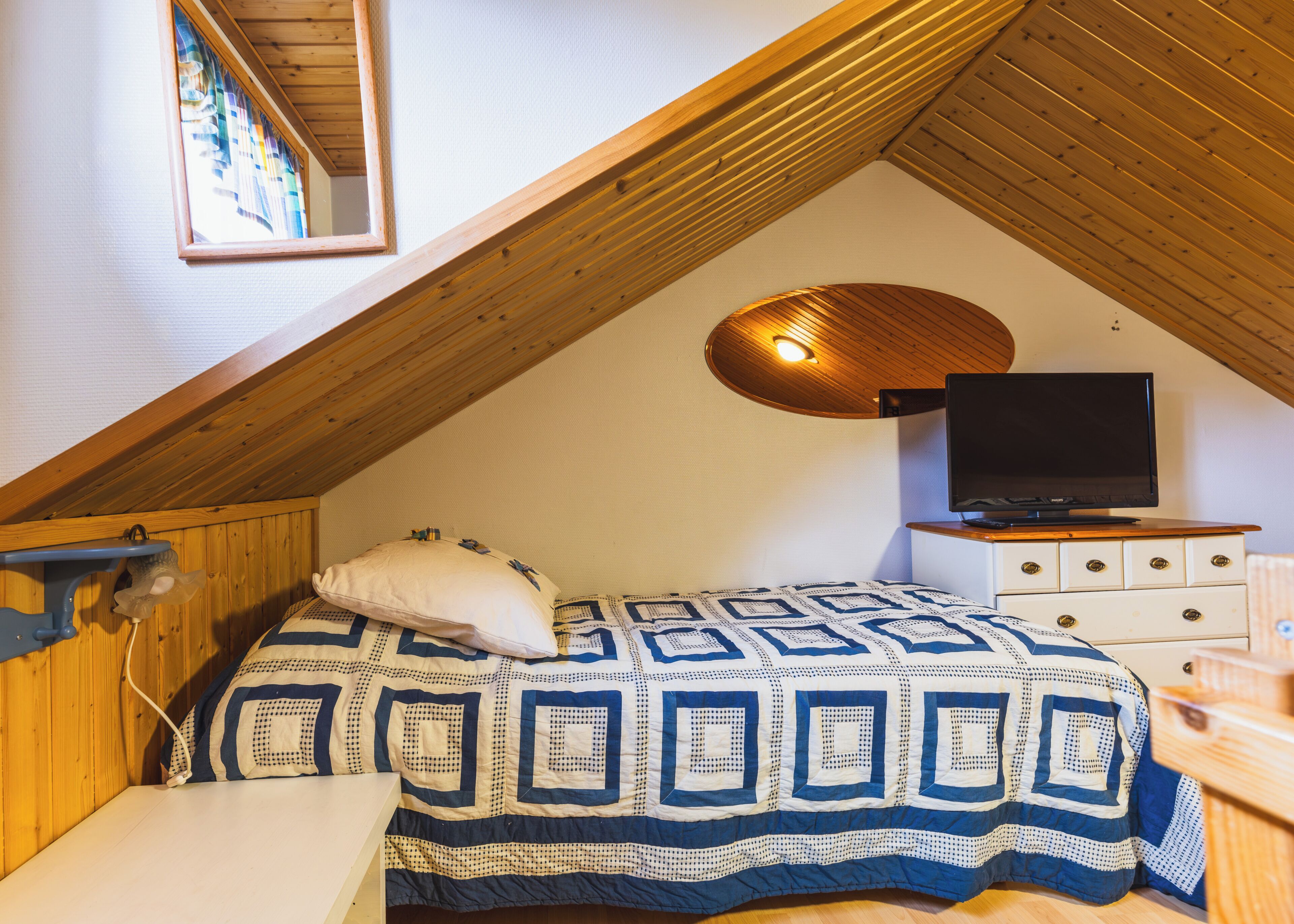 A bedroom with a sloped ceiling, a bed with a patterned bedspread, a television on a dresser, and a wall-mounted light fixture.