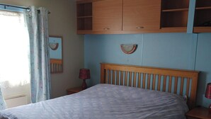 A bedroom with a bed, wooden headboard, a nightstand with a lamp, a mirror, and a wooden wardrobe. - Donegal Mobile Home-coast-family Friendly-sleeps 5 (Gortahork)