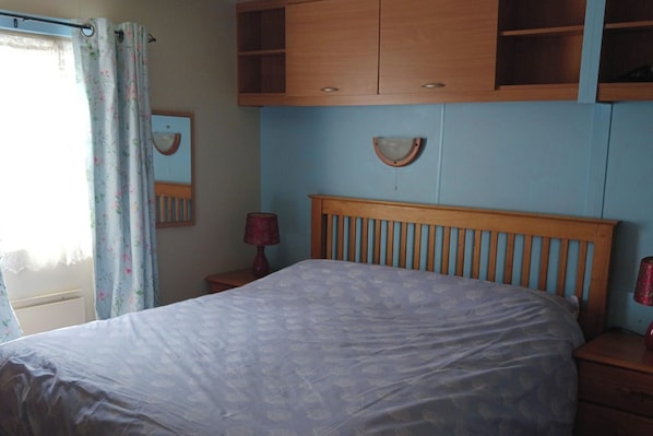A bedroom with a bed, wooden headboard, a nightstand with a lamp, a mirror, and a wooden wardrobe.