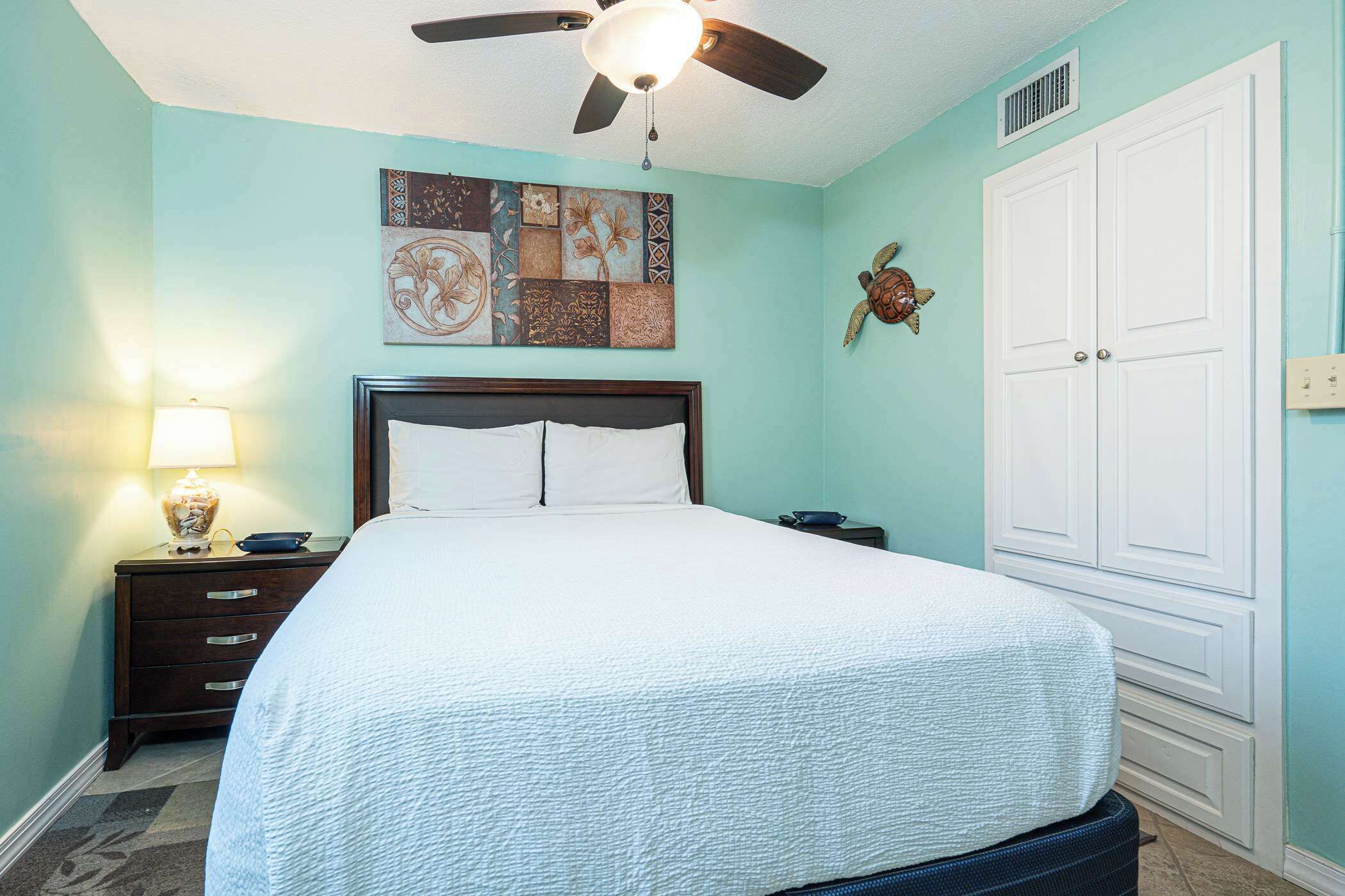 A bedroom with a bed, a nightstand, a lamp, a ceiling fan, and a wall decoration.