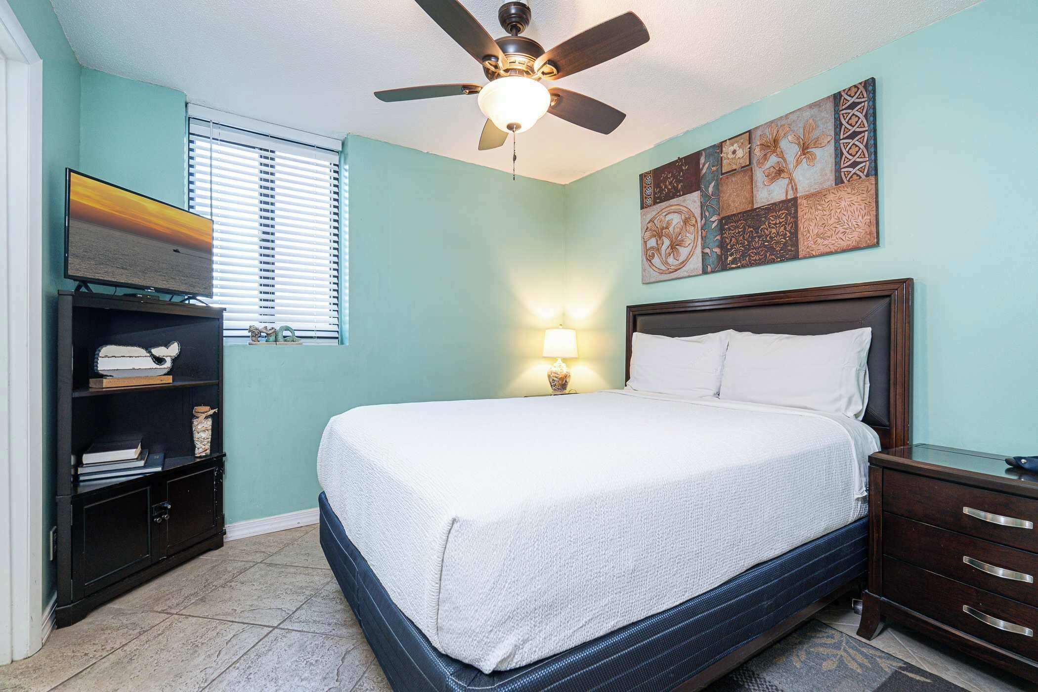 A bedroom with a bed, a ceiling fan, a TV, a lamp, and artwork on the wall.