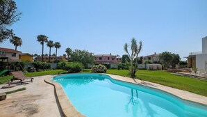 Uma piscina com borda curva, rodeada por um deck de concreto, e uma área gramada com árvores e um edifício ao fundo.