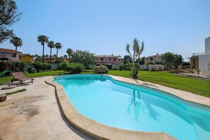 Ein Pool mit geschwungener Kante, umgeben von einer Betonplattform und einer Rasenfläche mit Bäumen sowie einem Gebäude im Hintergrund.