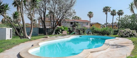 A swimming pool with a curved edge, surrounded by a concrete deck. There are palm trees and a building with a red roof in the background.