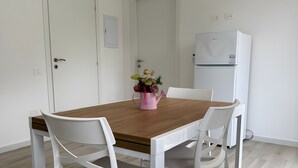 Uma área de refeições com mesa de madeira e cadeiras brancas, um refrigerador e um ar condicionado.