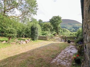 A stone pathway leading to a wooden gate, surrounded by lush greenery and a mountainous backdrop. - Frondirion (Machynlleth)