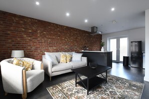 A modern living room with a brick wall, a sofa, a coffee table, and a kitchen area with a refrigerator. - Cozy corner - Perfect retreat only 10 minutes Walk to the Marina and Old Town (Kingston upon Hull)
