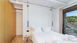Ein modernes Schlafzimmer mit einem großen Bett, einem Nachttisch, einem kleinen Hocker und Blick auf eine steinerne Treppe, die nach draußen führt.