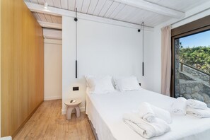 Ein modernes Schlafzimmer mit einem großen Bett, einem Nachttisch, einem kleinen Hocker und Blick auf eine steinerne Treppe, die nach draußen führt.