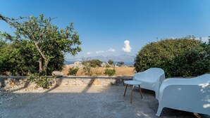 Ein Sitzbereich im Freien mit zwei weißen Stühlen, einer Steinmauer und Blick auf das Meer und die Berge.