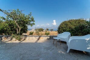 Ein Sitzbereich im Freien mit zwei weißen Stühlen, einer Steinmauer und Blick auf das Meer und die Berge.