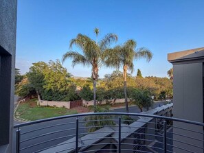 A balcony with a metal railing overlooking a garden with palm trees and a clear sky.