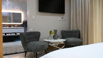 Habitación de hotel con cama, dos sillones, una mesita redonda, televisor de pantalla plana en la pared y un ventanal con cortinas.