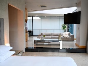 A modern hotel room with a large bed, a glass coffee table, a sofa, and a flat-screen TV mounted on the wall.