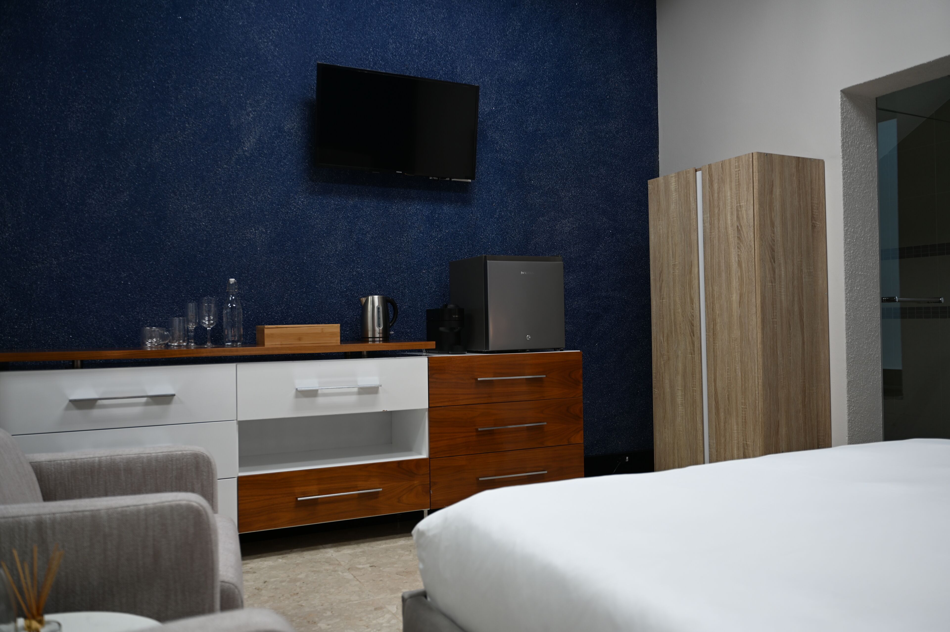 Un dormitorio moderno con cama, cómoda de madera, televisor de pantalla plana en la pared y mesita de noche con lámpara.