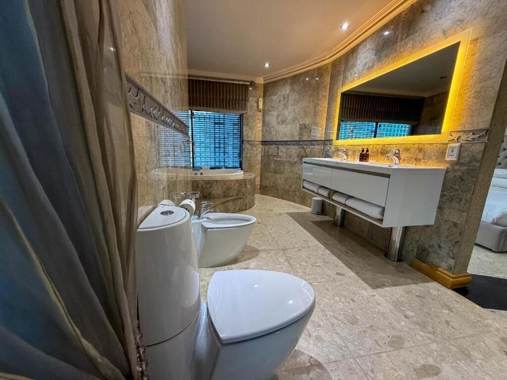Baño con inodoro, bidé y lavamanos, paredes de mármol y un espejo grande.