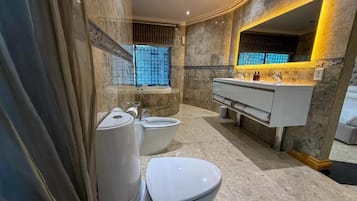 Baño con inodoro, bidé y lavamanos, paredes de mármol y un espejo grande.