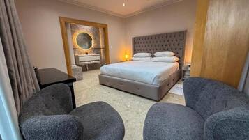 A bedroom with a large bed, two armchairs, a desk, and a mirror with a decorative frame.