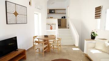 A modern living room with a flat-screen TV, a round dining table with chairs, a sofa, and a staircase leading to a kitchen area.