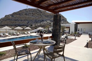 A pool area with lounge chairs and umbrellas, a table with a vase and wine glasses, and a view of a mountainous landscape. - THERRA Suites & Sunset Terrace (OÃa)