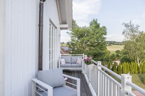 A balcony with a grey sofa and cushions, a white railing, and a view of a green landscape with trees and a field.
