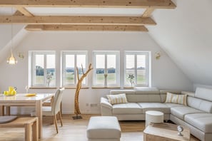Un soggiorno moderno con soffitto in legno, un divano bianco, un tavolo da pranzo con sedie e ampie finestre vista sulla campagna.