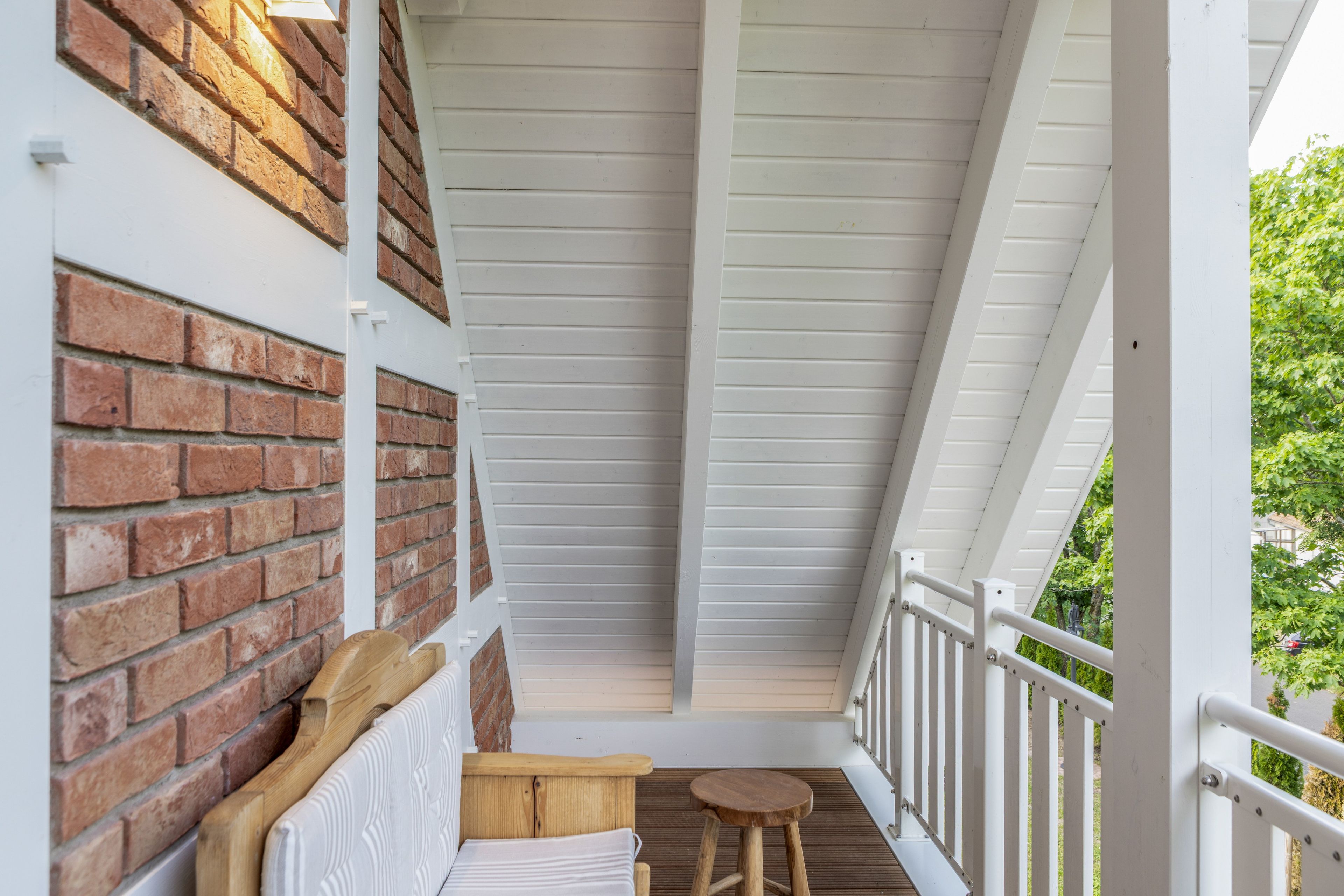 Een overdekt balkon met een bakstenen muur, een houten bankje, een klein krukje en een witte balustrade.