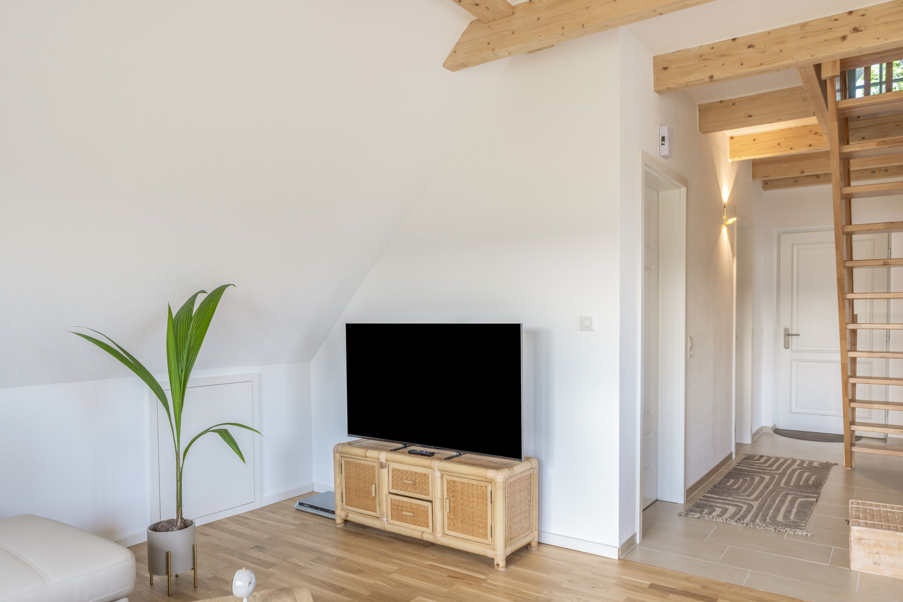 Una sala de estar con un televisor sobre un mueble de madera, una planta en maceta y una escalera que conduce a otra habitación.