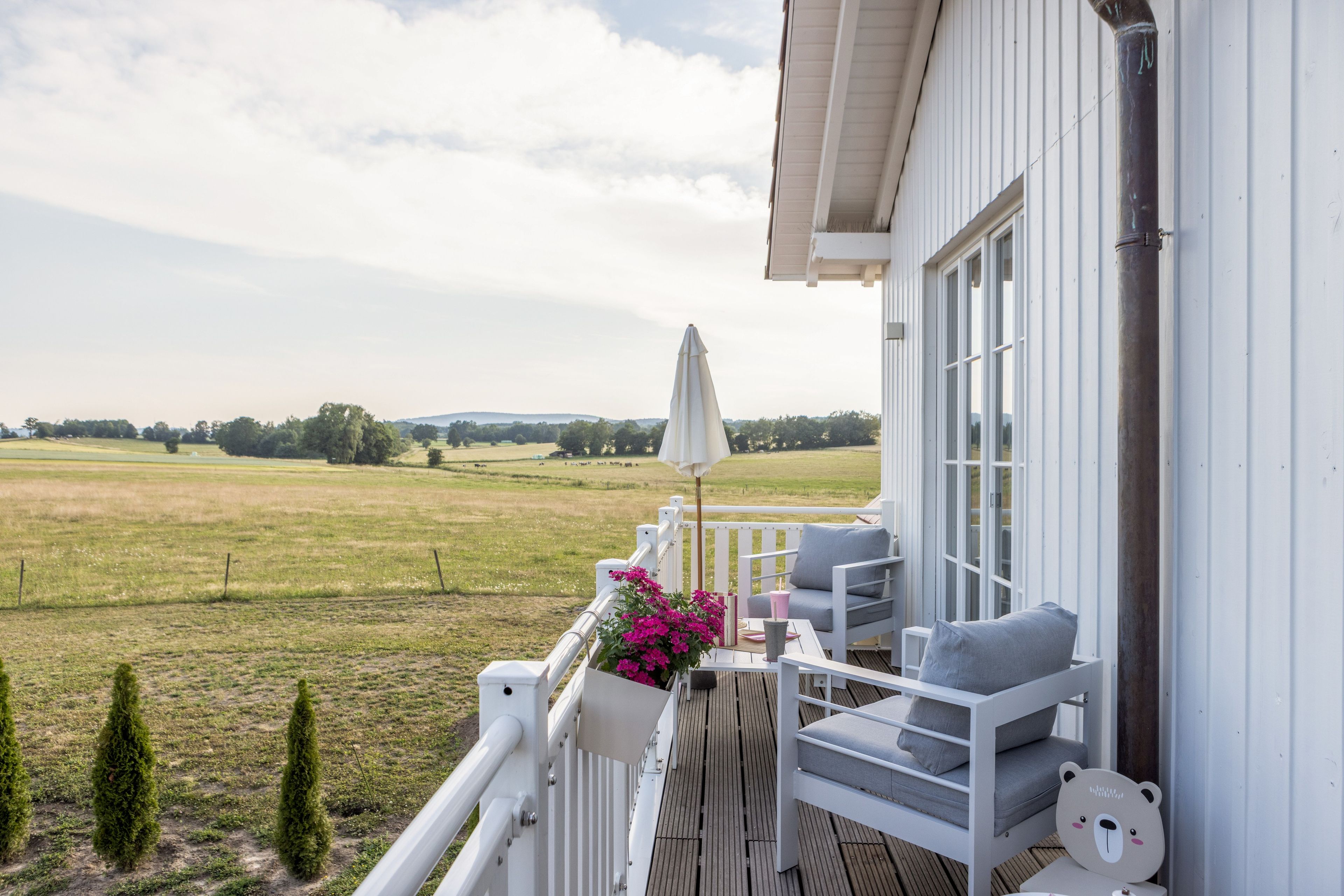Ein Balkon mit Blick auf ein grünes Feld, einem weißen Geländer sowie einem Tisch mit einem Stuhl und einem Blumentopf.