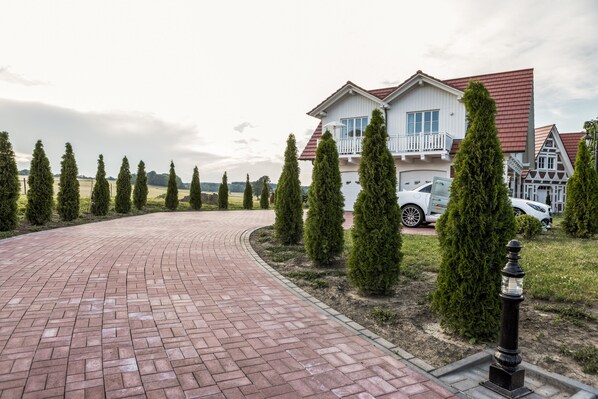 A paved driveway leading to a two-story house with a balcony, surrounded by neatly trimmed evergreen trees.