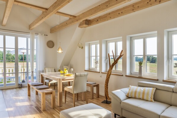 A spacious room with a dining area, a sofa, and large windows offering a view of the outdoors.