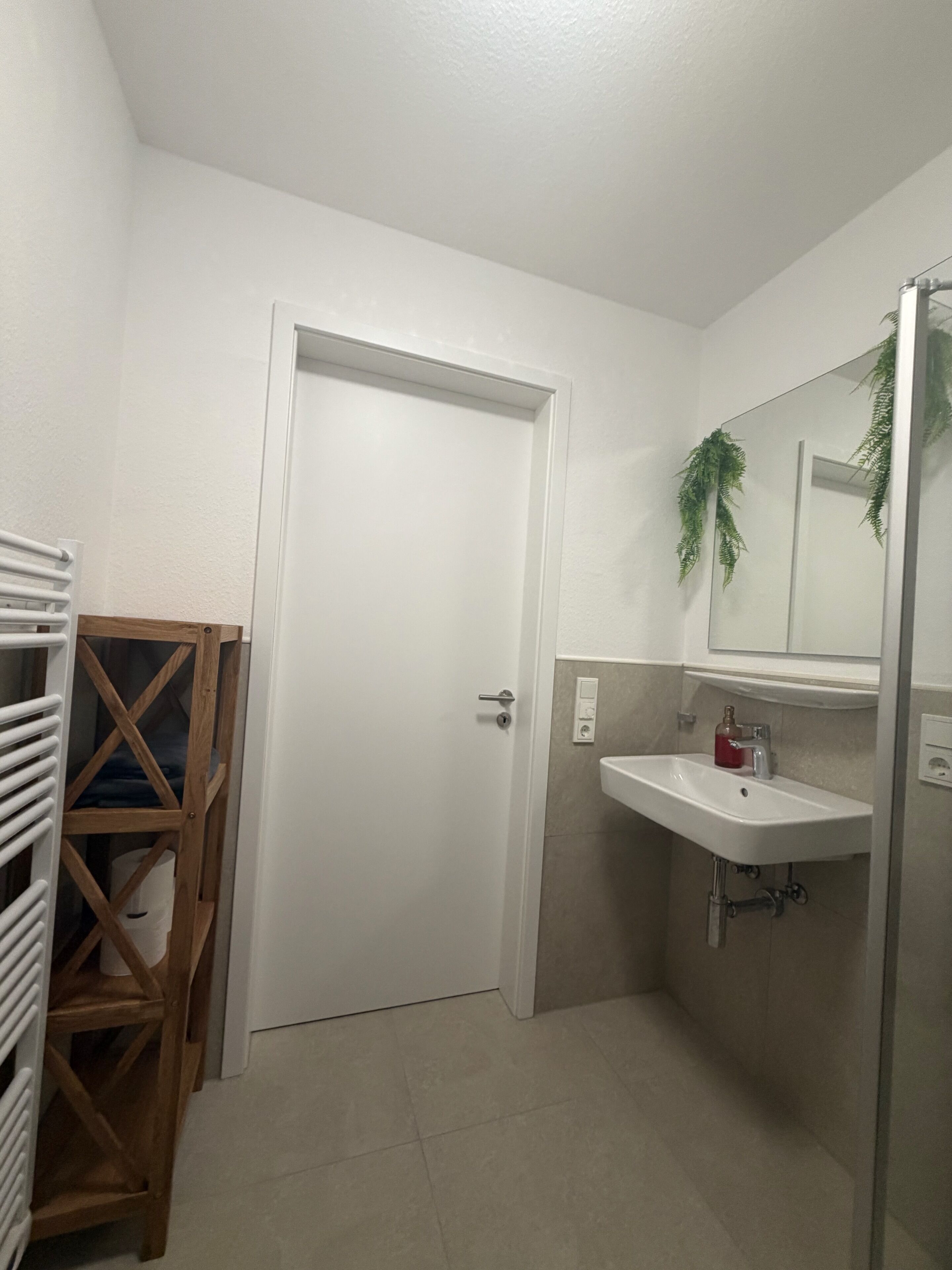 A bathroom with a white door, a wooden shelf, a sink, and a mirror.