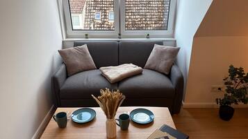 Ein gemütliches Zimmer mit einem grauen Sofa, zwei Kissen, einem kleinen Tisch mit einer Vase, Tellern und einem Buch.