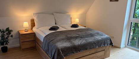 A bedroom with a wooden bed, two bedside tables with lamps, a potted plant, and a window with a view.