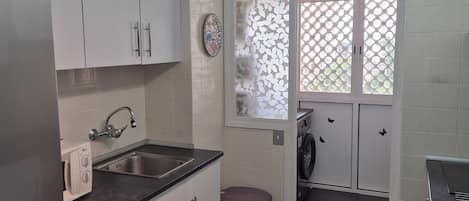 A compact kitchen with white cabinets, a stainless steel sink, a washing machine, and a clock on the wall.