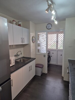 A compact kitchen with white cabinets, a stainless steel sink, a washing machine, and a clock on the wall. - Vacation Rental Apartment Near the Beach (VC)