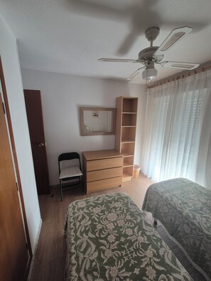A bedroom with a bed, a chair, a dresser, a shelf unit, and a ceiling fan. - Vacation Rental Apartment Near the Beach (VC)