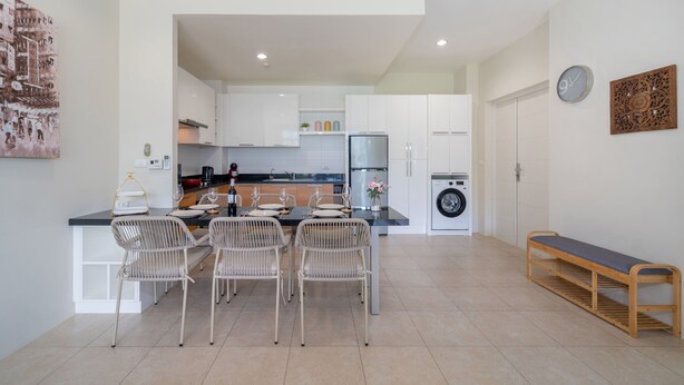 A modern kitchen with white cabinets, a dining area with wicker chairs, a built-in washing machine, and a bench.